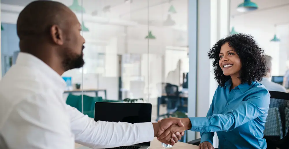 A man and woman shaking hands across a desk whilst smiling