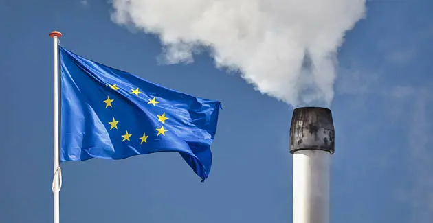 An EU flag next to a billowing factory chimney. The flag is a circle of 12 gold stars on a blue background 