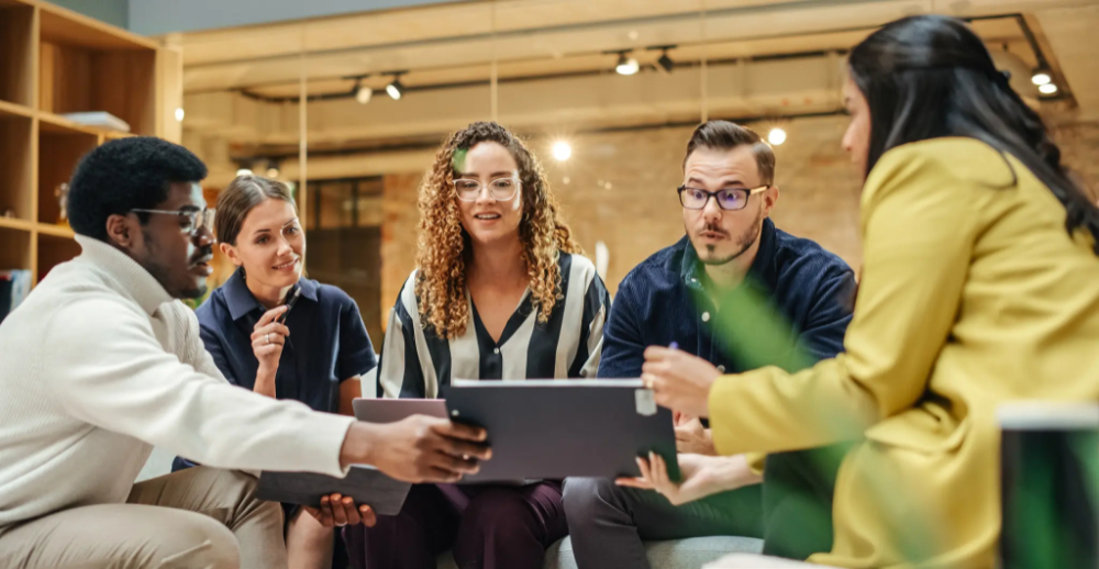 Five people sat grouped around an ipad being held between the two furthest outside people.  