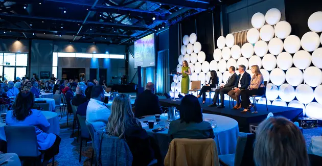 Conference panel discussion on stage with audience seated at round tables, professional event with speakers and attendees.