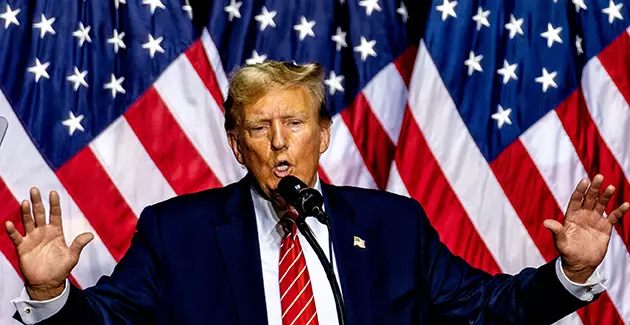 Man in a suit speaking at a podium with multiple American flags in the background.