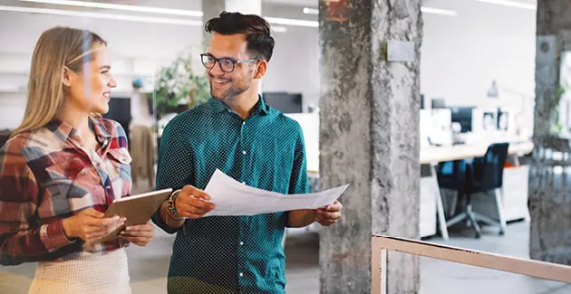 Man and woman in an office looking at each other, one holding an ipad and the other holding a piece of paper