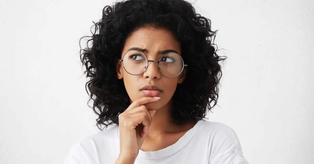 A woman with black curly hair, wearing glasses with her hand on her chin thoughtfully