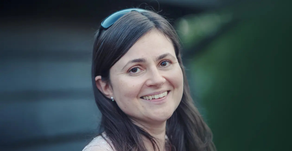 A woman with long brown hair, wearing sunglasses on her head, smiling at the camera