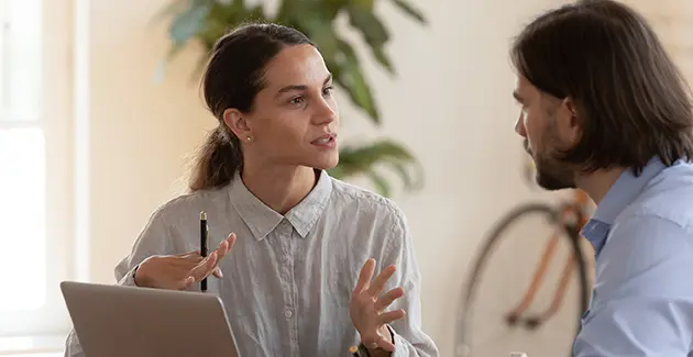 A woman with a laptop infront of her is looking at a man, they are both wearing shirts and have brown hair. The woman is gesturing with her hands and is also holding a pen. The man is looking at her. 