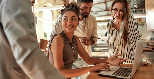 Smiling people standing around a woman sat at a laptop