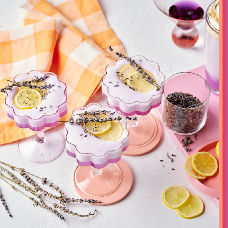 Three flower shaped cocktail glasses containing purple hued cocktail on white backdrop with cocktail shaker, lavender and lemon.