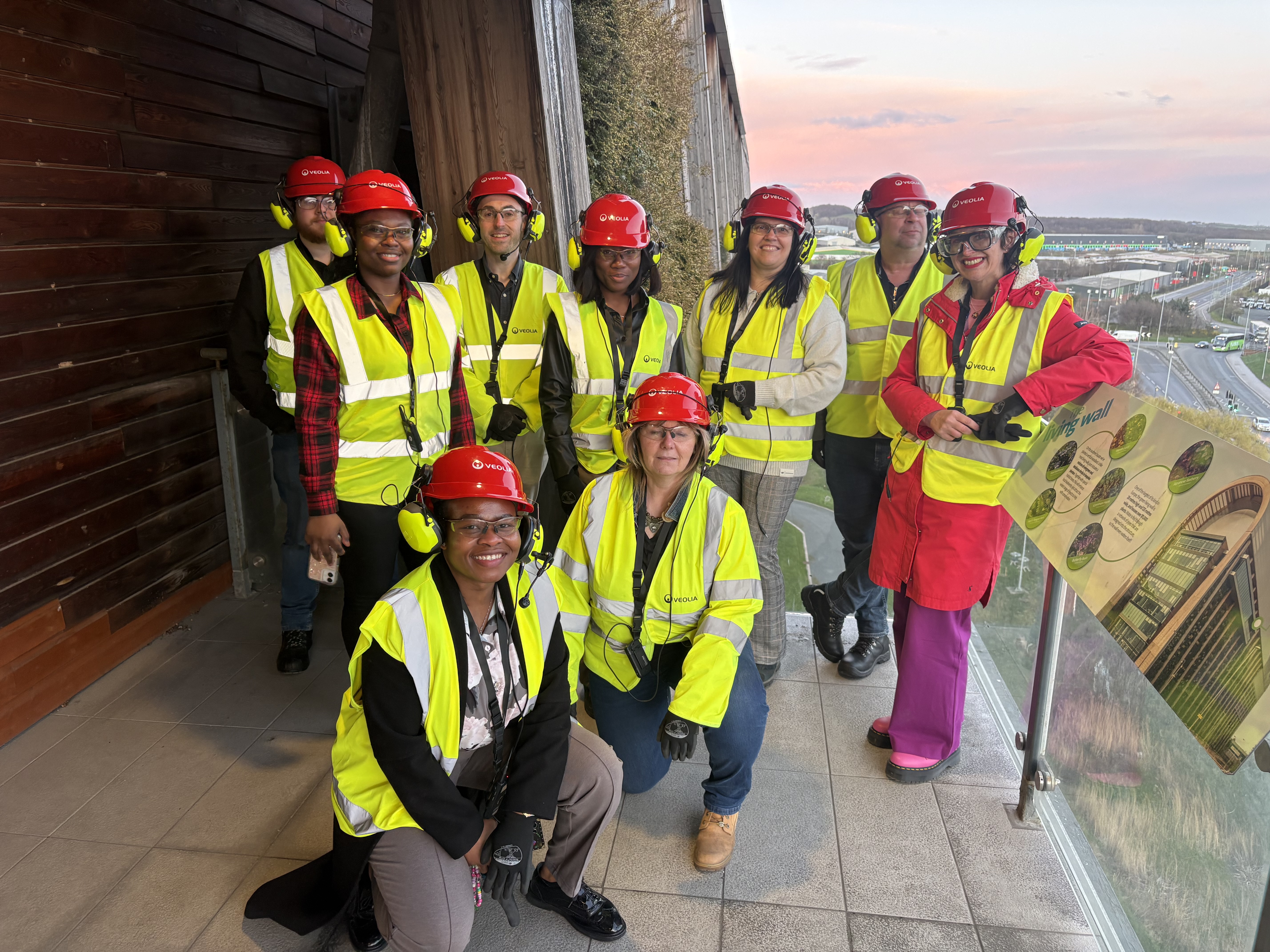 A group of nine people wearing red hard hats, yellow high-visibility vests, and ear protection standing on an outdoor balcony overlooking a road.