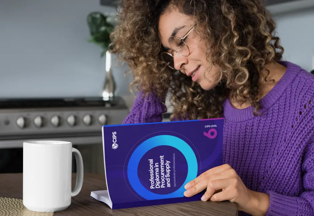 A woman with curly hair and glasses, wearing a purple jumper, sitting at a table and reading a book titled Professional Diploma in Procurement and Supply with CIPS LEVEL 6 branding. 