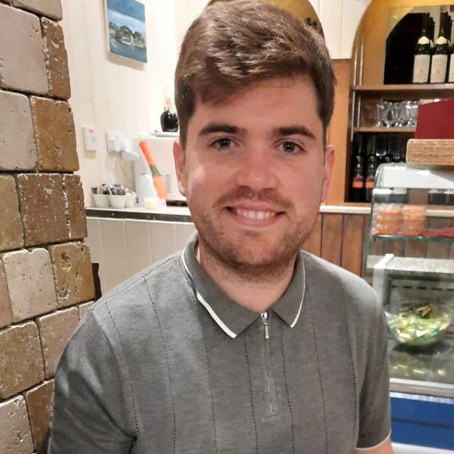 A casual indoor photograph of a smiling young man with brown hair. He is wearing a grey textured polo shirt and is seated in a space with a light-colored brick wall on the left and a bar or service area in the background.
