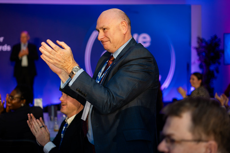 A man in a blue suit and patterned tie stands and claps his hands in a crowded room with blue lighting.