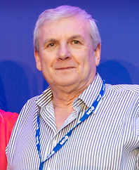 A portrait of a man with short grey hair wearing a blue and white striped button-down shirt and a blue lanyard.