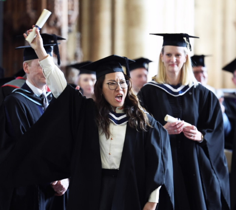 A jubilant graduate in cap and gown raises a diploma scroll in triumph, surrounded by other graduates.