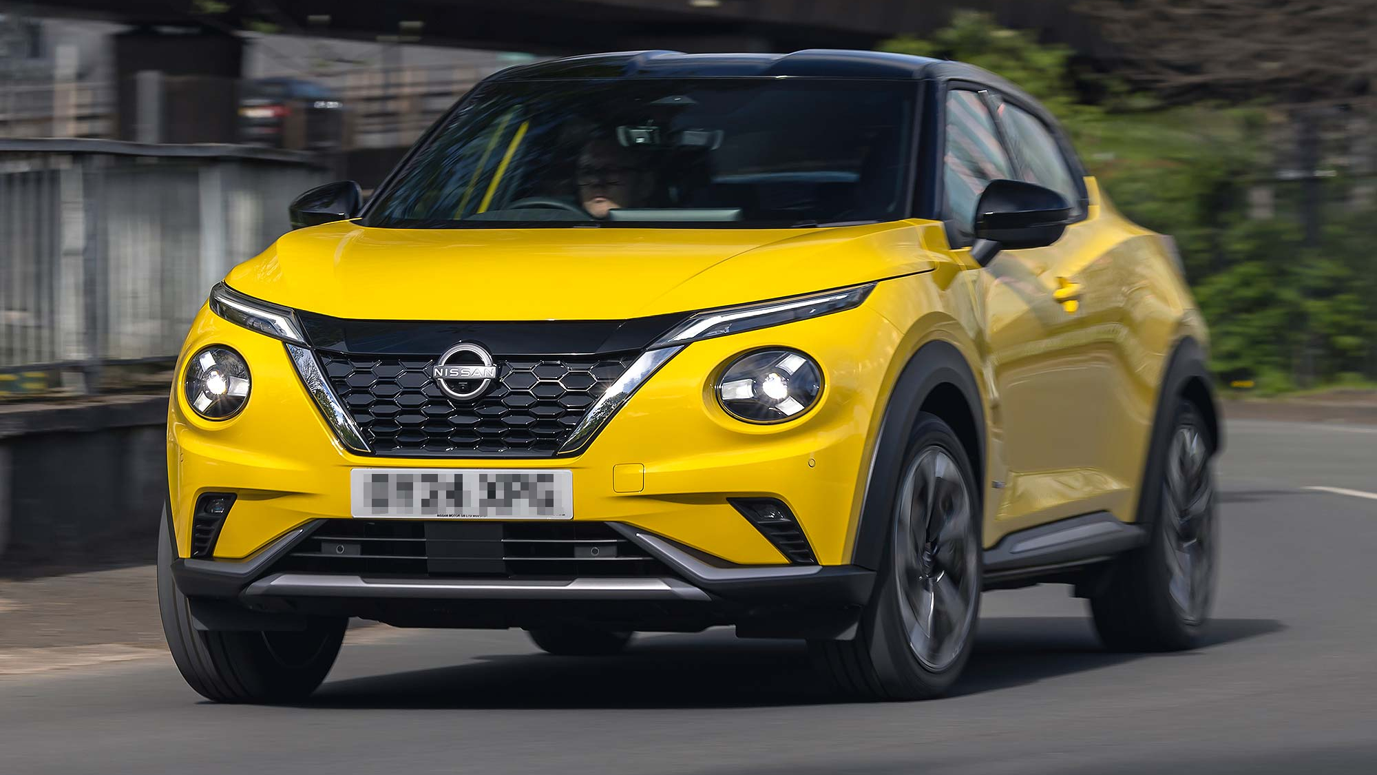 A bright yellow Nissan Juke compact SUV with black roof detailing is driving on a city road.