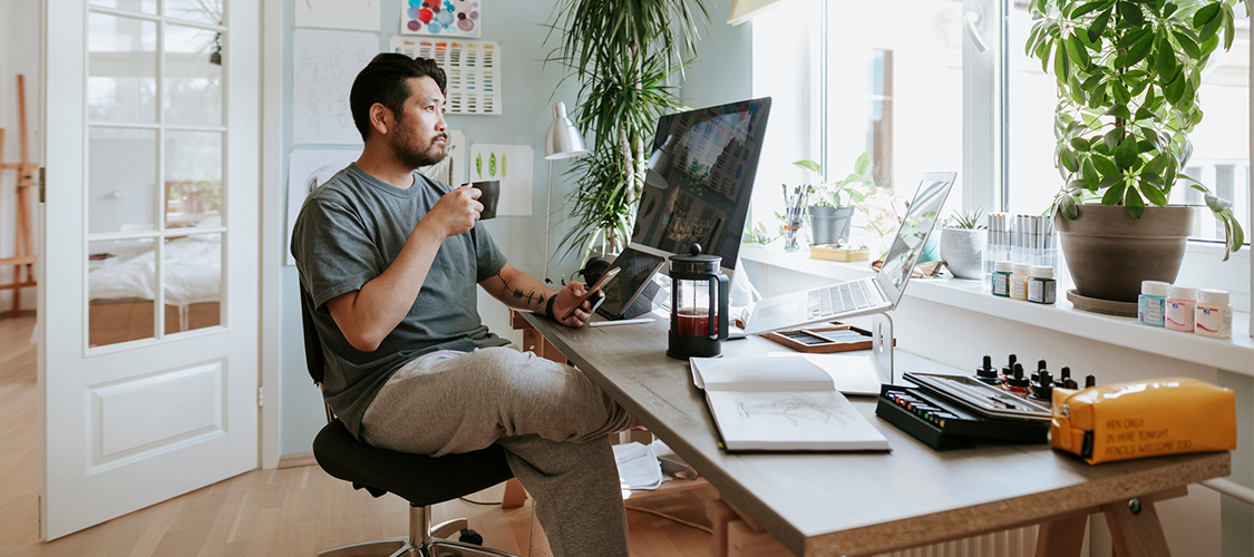A man sitting in a home office working on a computer and drinking coffee.