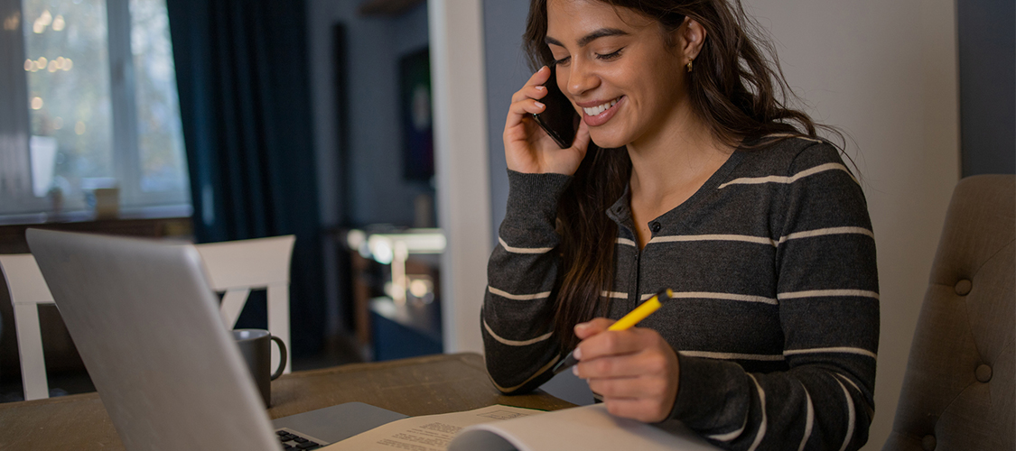 A smiling woman looking at a laptop as she writers in a notebook.