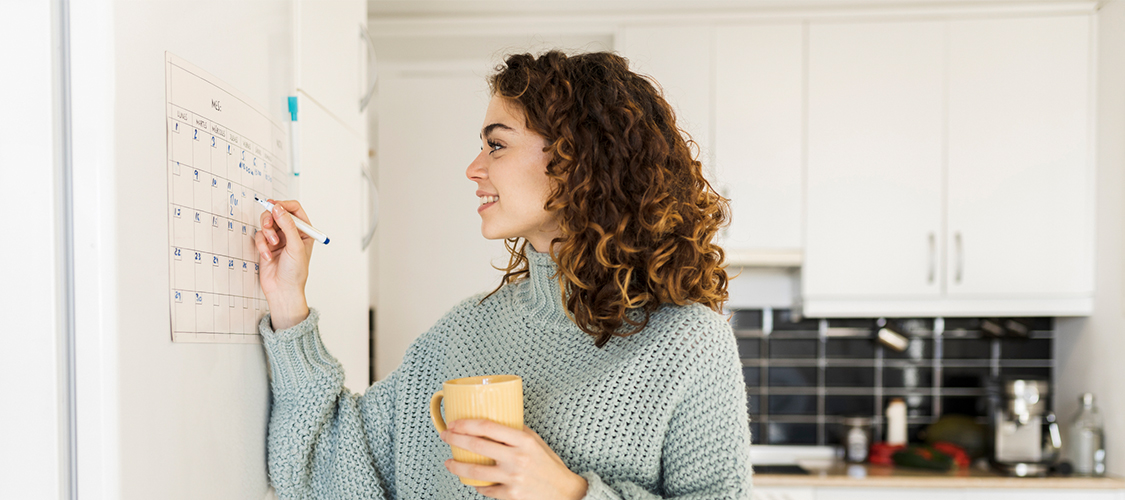 A woman marking off a date from a large calendar hanging in a kitchen.