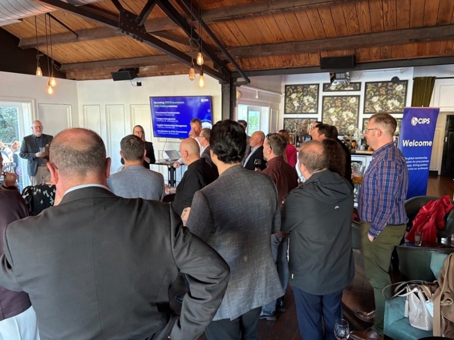 A large group of approximately 20 people, dressed in business and smart casual attire, posing for a group photo on a stage. They are standing in front of a blue CIPS UK & Europe Volunteer Conference and Awards 2025 banner. Several individuals in the center are holding awards.