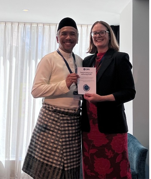 A man wearing traditional Malay attire (white shirt, songkok, and plaid sarong) posing for a photo with a woman in a red dress and dark blazer. The woman is holding a CIPS certificate that says 'Congratulations on Renewing FCIPS.' Both are smiling.