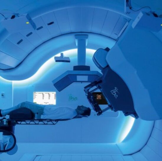 A PROTEUS ONE proton therapy unit supplied by IBA is shown hovering over a patient and preparing for a scan.