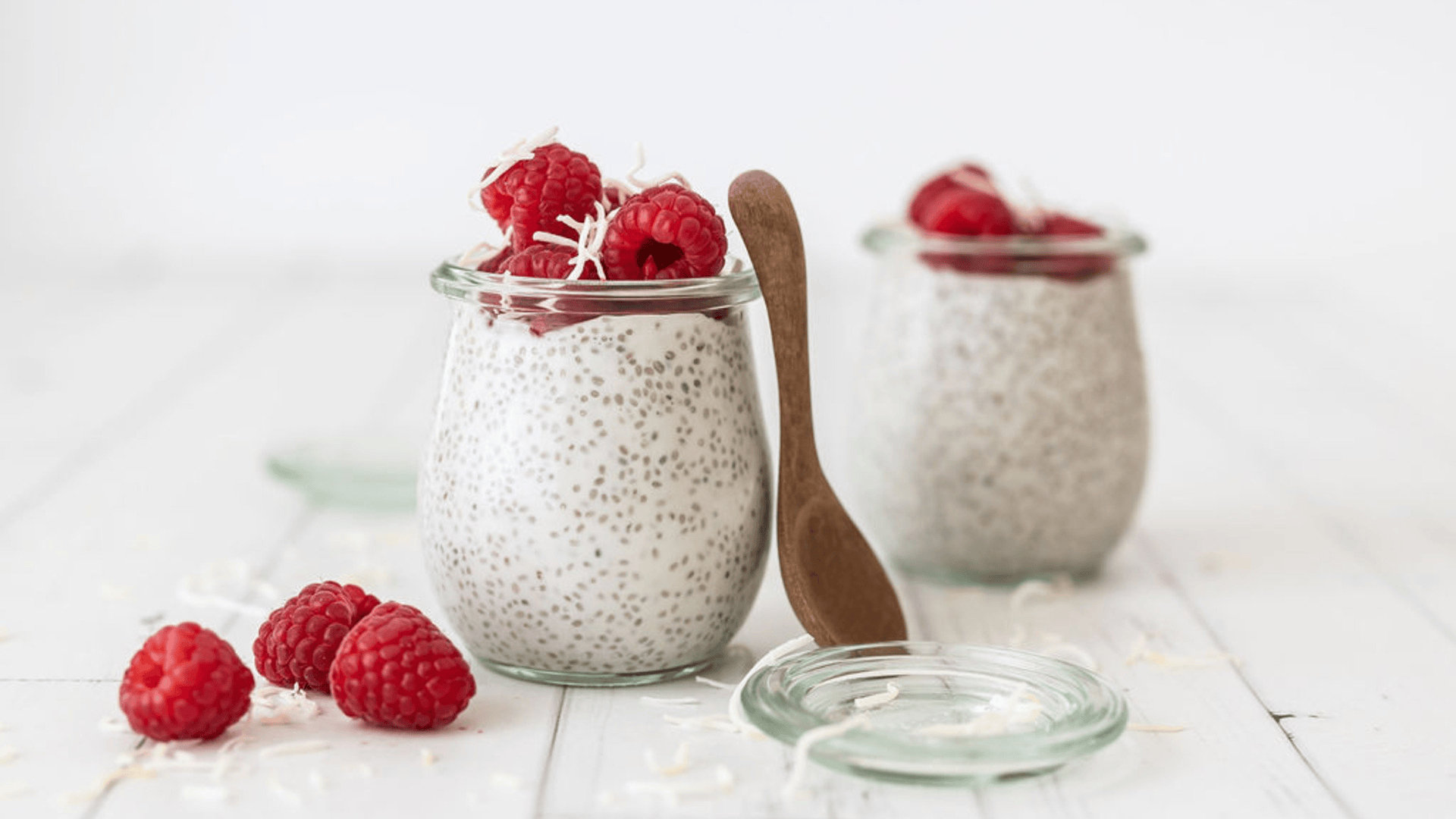 Two chia puddings in glass jars are featured on a white wooden surface. The chia puddings are topped with fresh raspberries and sprinkled with shredded coconut.
