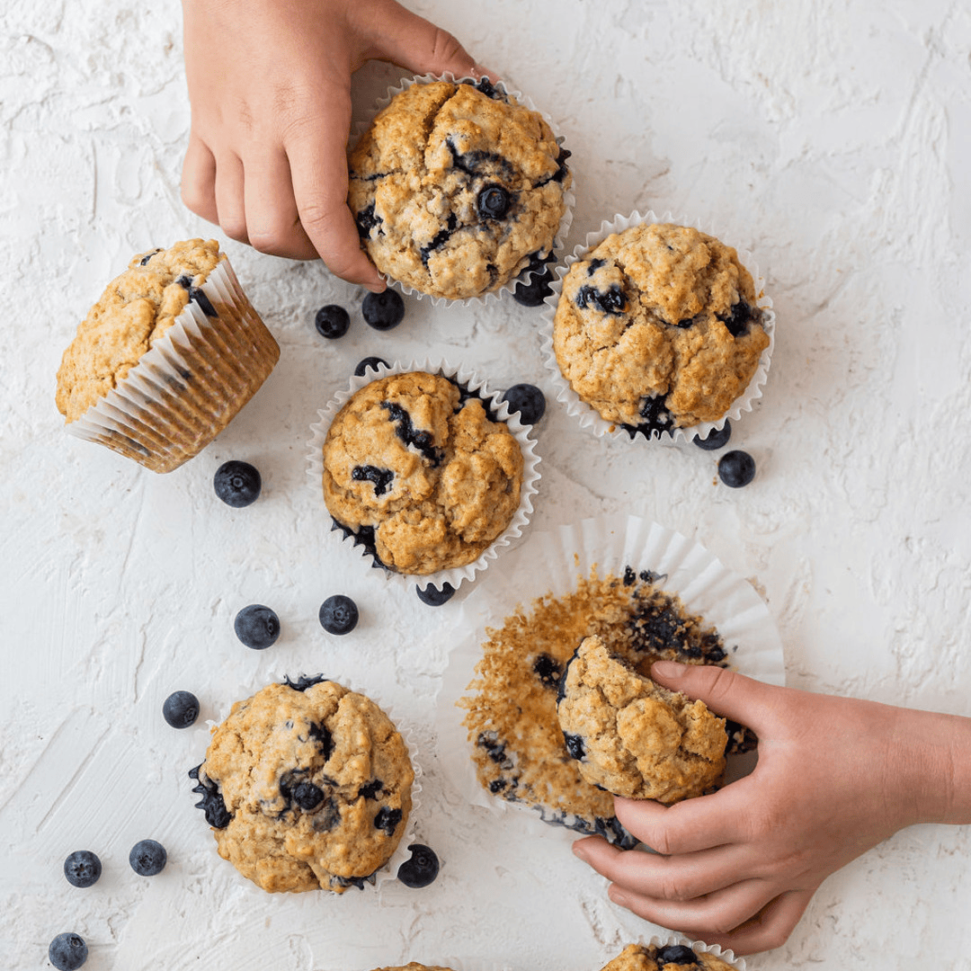 Nine homemade blueberry muffins are spread across a white kitchen benchtop, amongst scattered blueberries. Two sets of children’s hands are reaching for the muffins.