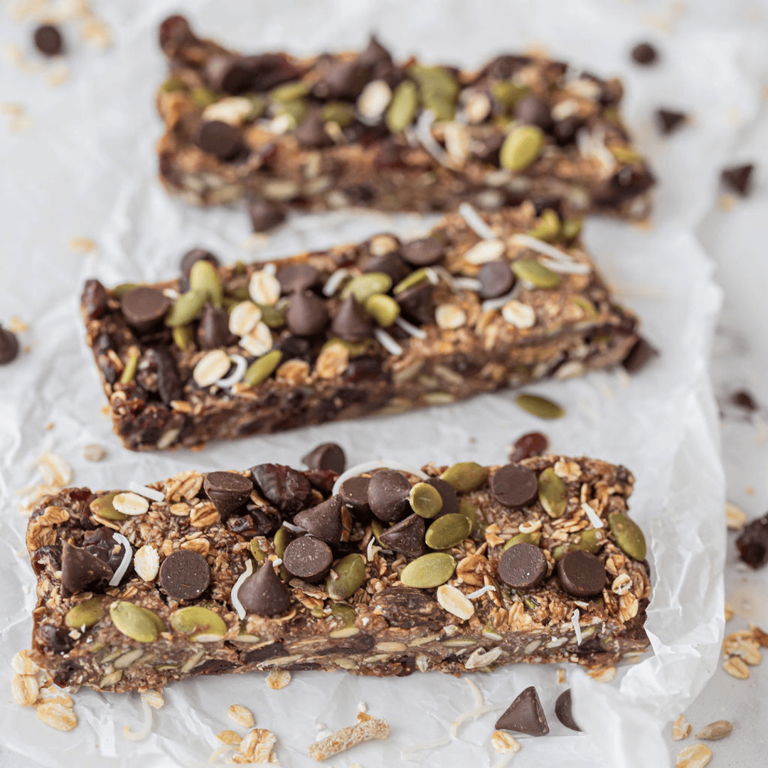 Three homemade granola bars are placed upon baking paper. The bars are sprinkled with pepitas, dark chocolate drops and shredded coconut.