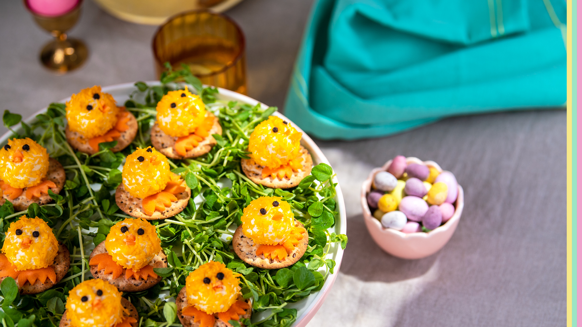 Mini cheese balls rolled in shredded cheese and decorated to look like baby chicks sitting on a bed of herbs with sugar Easter eggs sitting behind 