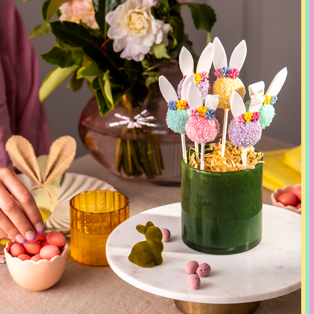 Multi coloured Easter bunny cake pops rolled in desiccated coconut displayed in dark green vase with sugar Easter eggs in background