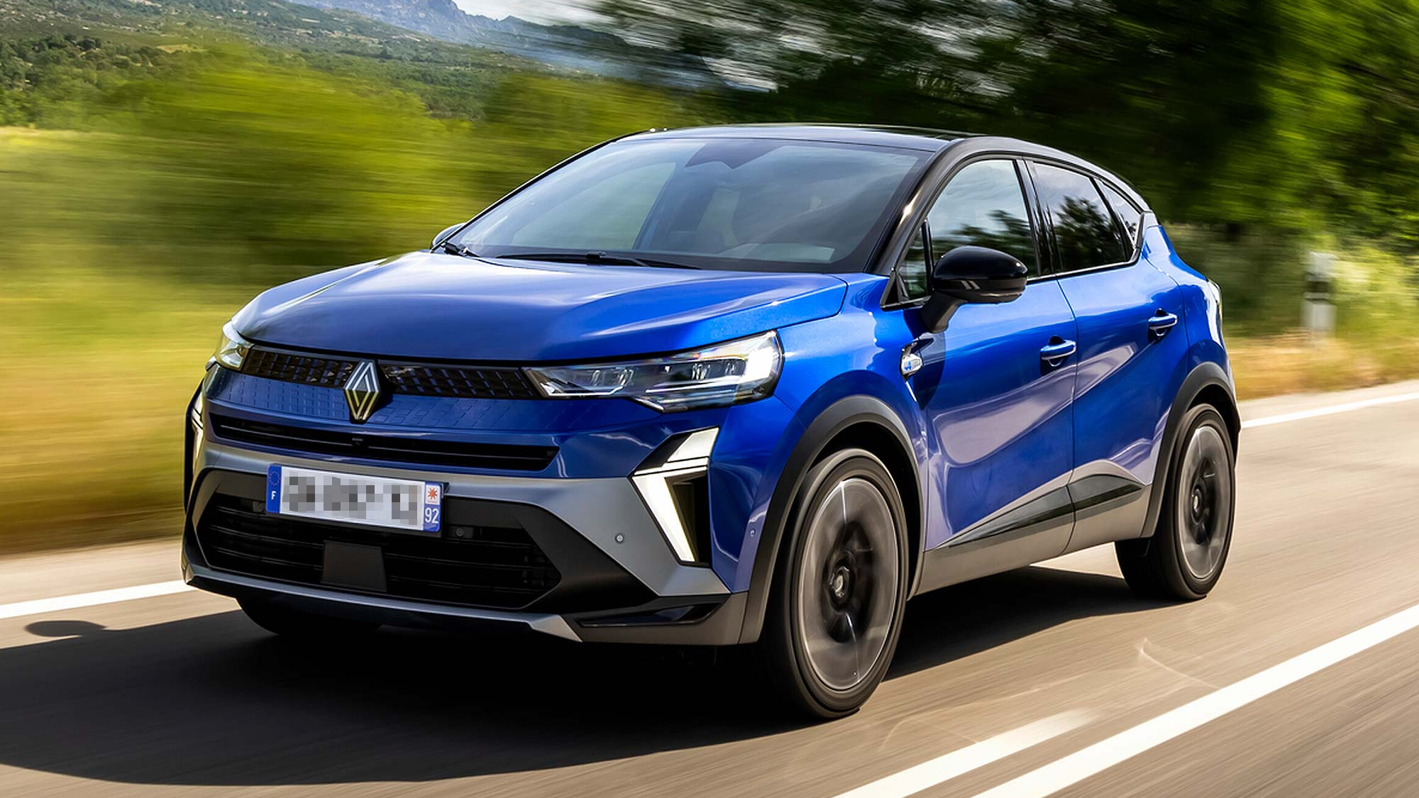 Blue Renault Captur SUV driving along a country road, with blurred trees and mountains in the background.Blue Renault Captur SUV driving along a country road, with blurred trees and mountains in the background.