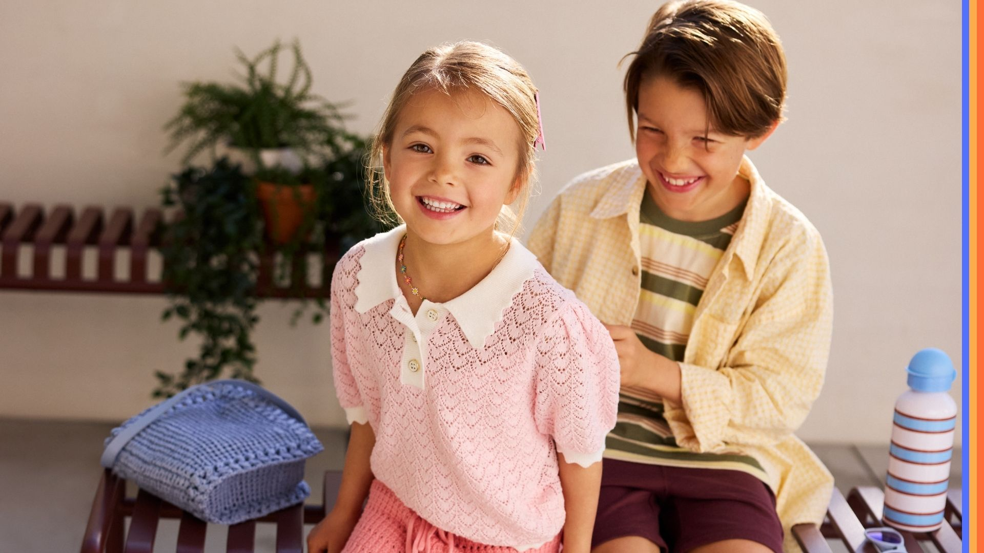 Two children sitting side by side outdoors, each holding a colorful water bottle. The little girl wears a light pink top with a ponytail, and the boy wears a yellow shirt over a stripey t-shirt.
