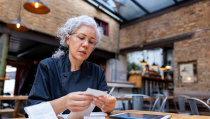 Female business owner looking over receipts at a table while using a tablet 