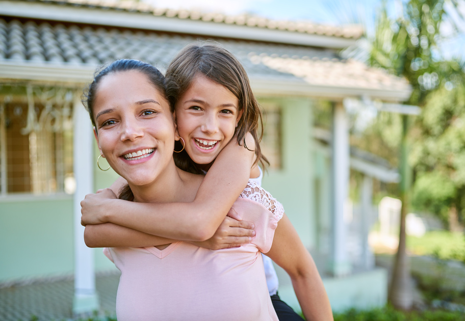 Smiling woman giving a piggyback ride to a young girl outside a green house on a sunny day.