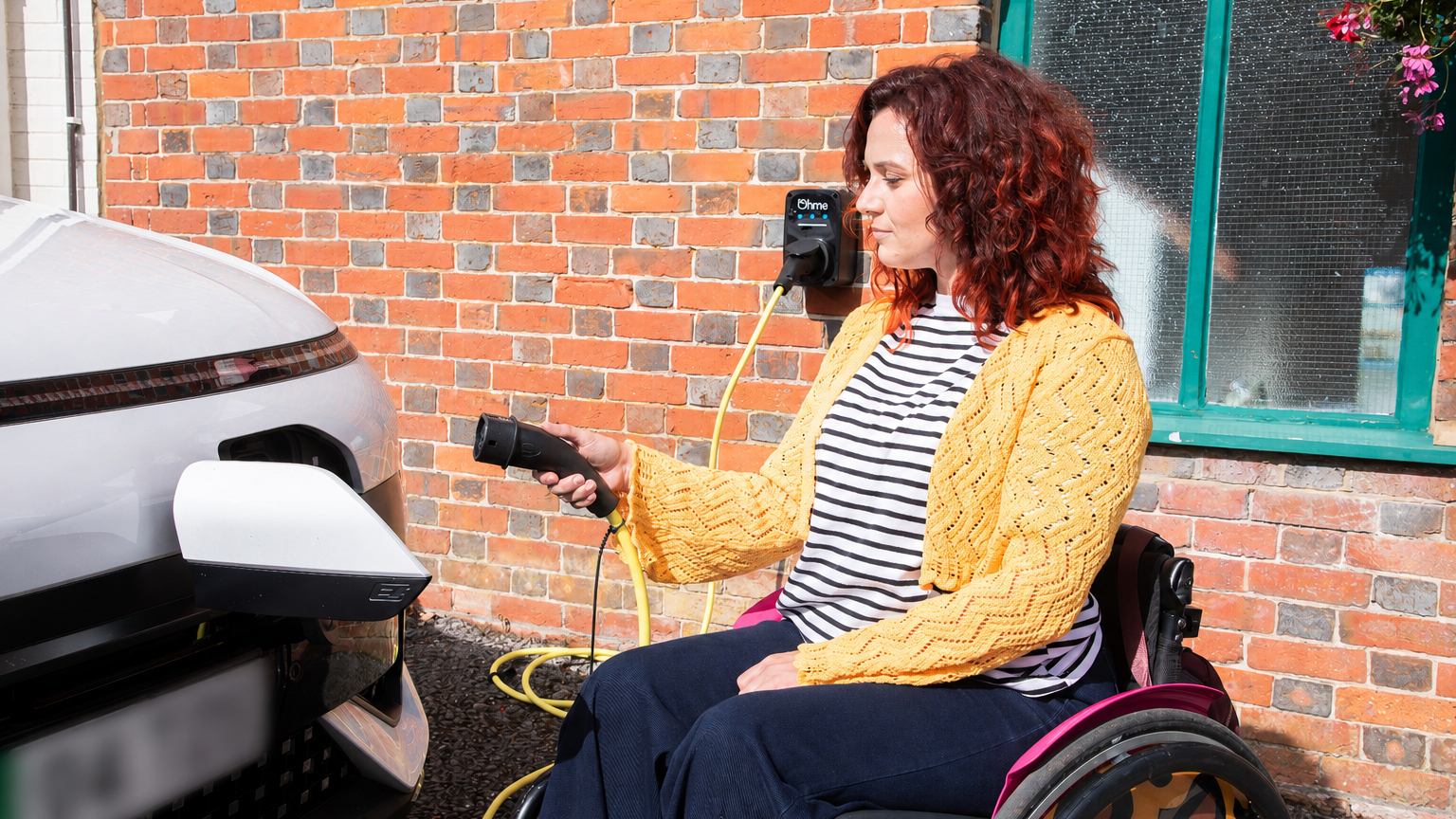 A woman using a wheelchair plugs an electric vehicle into a home charger mounted on a brick wall, holding the charging cable beside a light-coloured car.