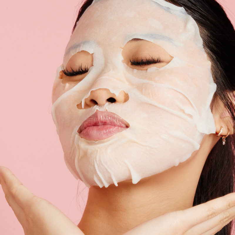 Woman wearing a hydrating H2O+ Vitamin C Glow Face Mask with eyes closed on a pink background.