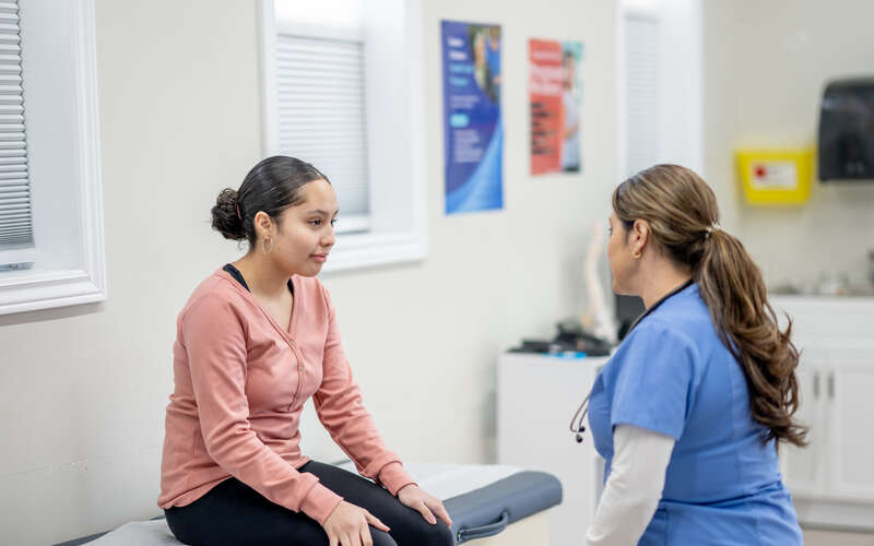 A young female teenager sits up on an exam table at the doctors during a routine check-up. She is dressed casually and her female nurse of Hispanic decent, is seated in front of her as they talk.