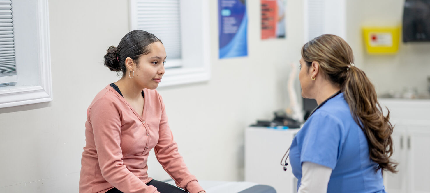 A young female teenager sits up on an exam table at the doctors during a routine check-up. She is dressed casually and her female nurse of Hispanic decent, is seated in front of her as they talk.