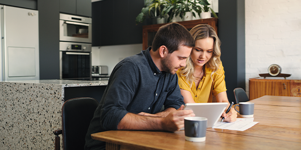A white man and woman sit at a table looking at a tablet and writing on a piece of paper