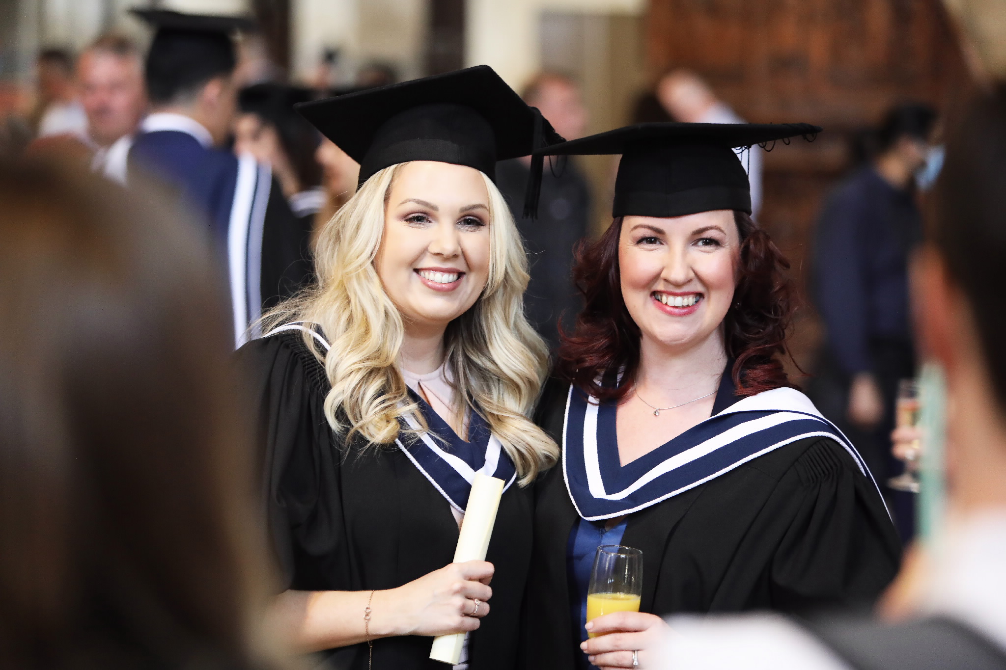 Two female graduates smiling with a rolled diploma and drink