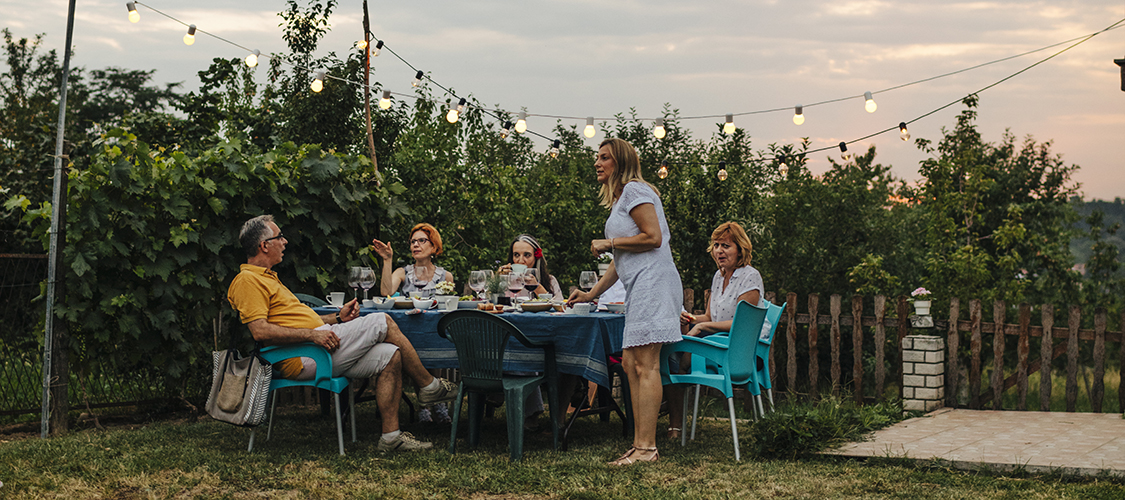 A group of adults hang out around a table under string lights outside of a home