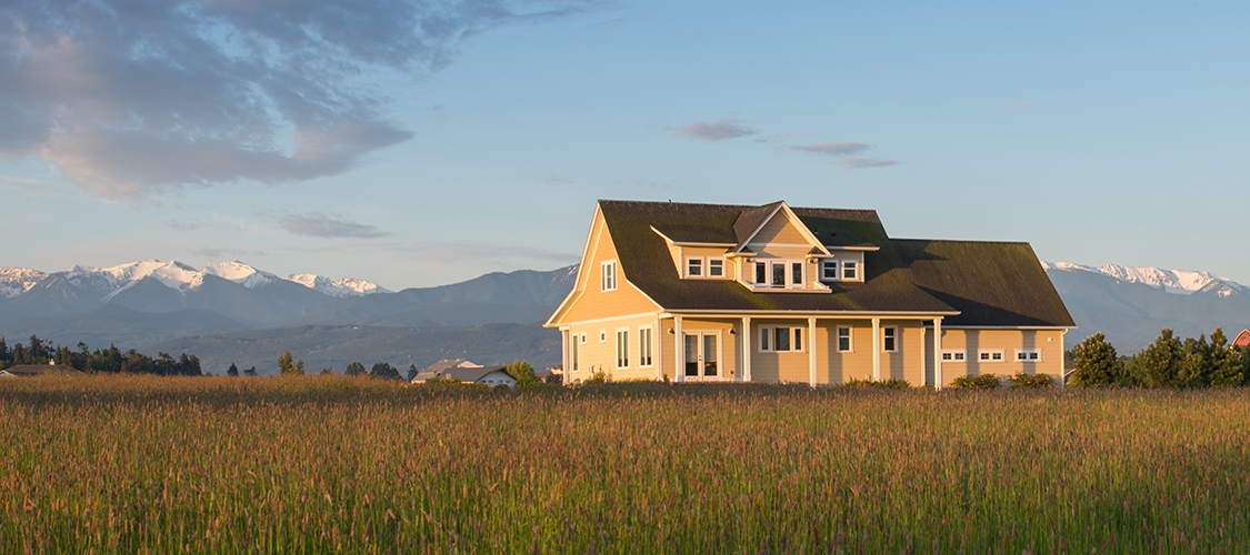 A large two story house sits on open grass land with mountains in the background