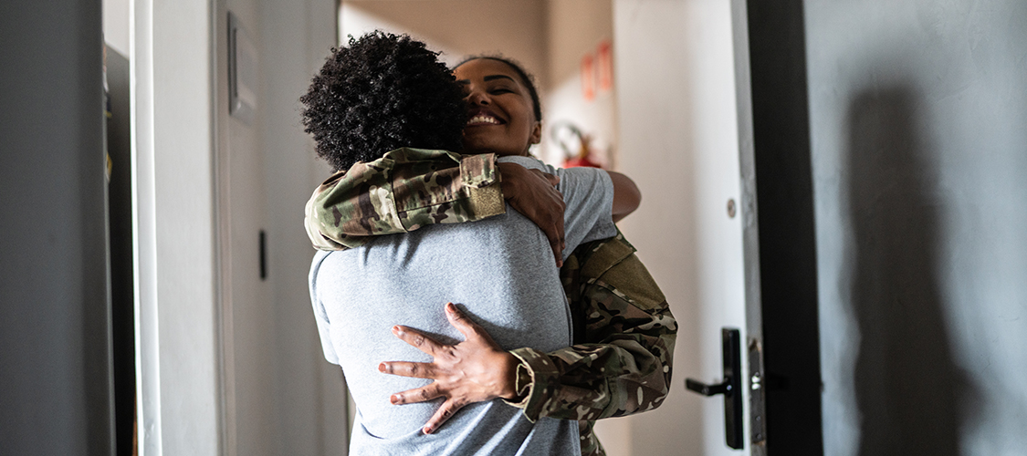 A Black woman in a military uniform smiles while she hugs another person