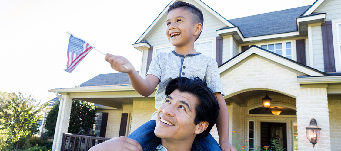 A Hispanic man stands in his front yard with a little boy holding an American flag on his shoulders