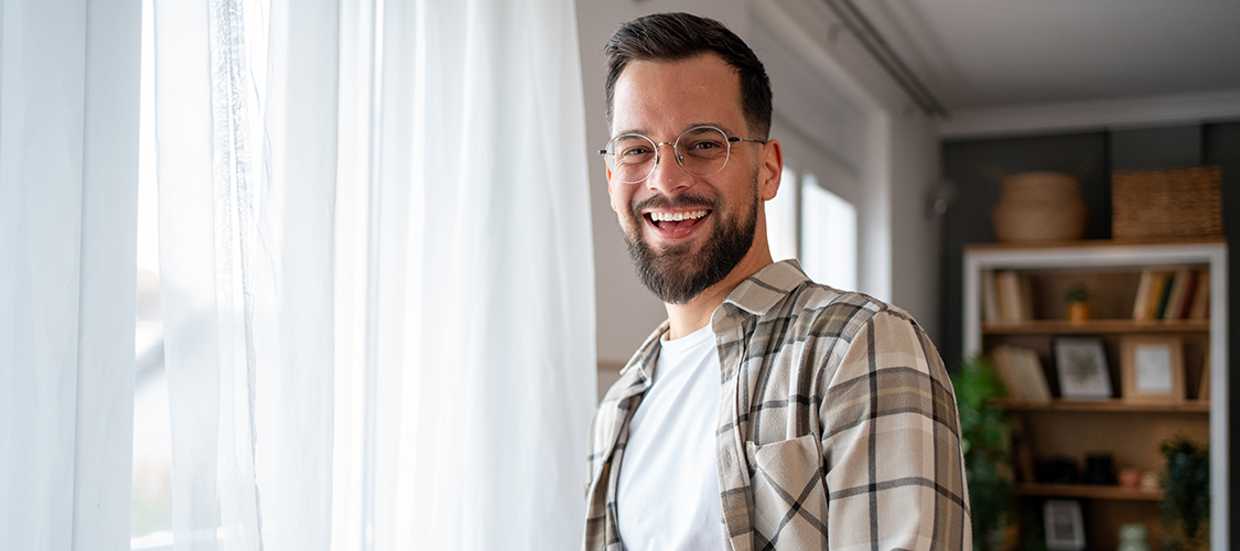 A white man stands smiling beside the window of his home