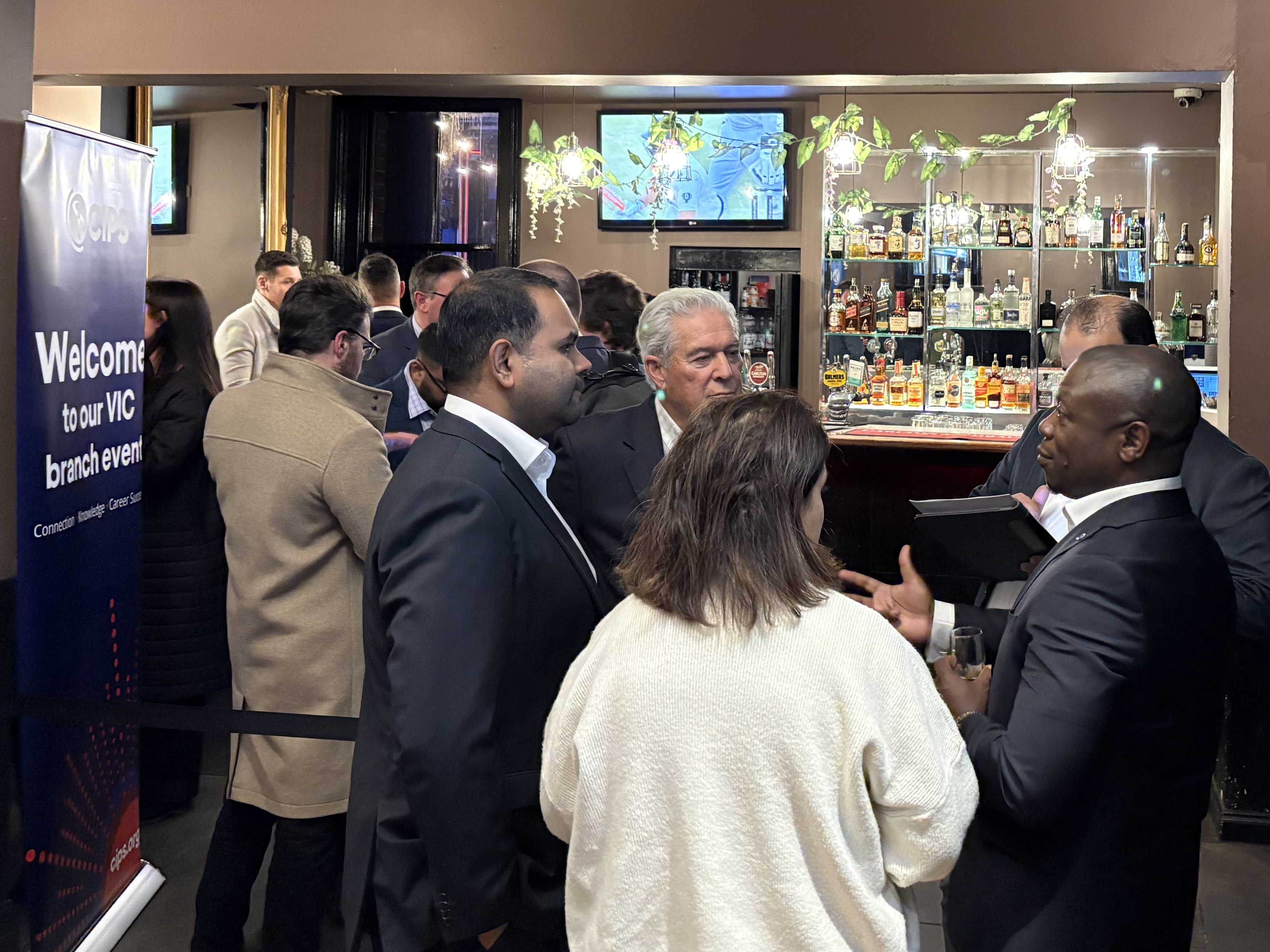 A group of people networking at an evening event, standing around a bar area. A CIPS banner is visible on the left. The scene shows a professional networking event.