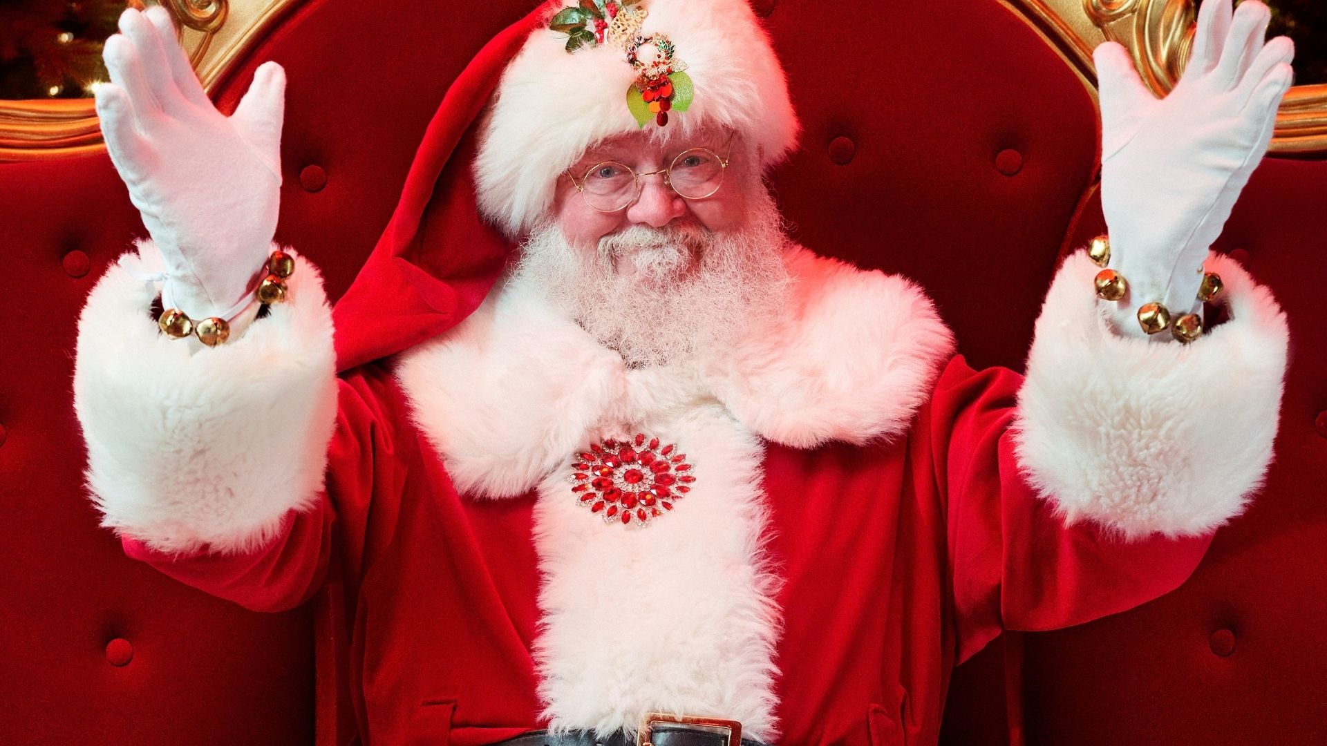 Santa raises both gloved hands in a wave from his red velvet and gold throne at [Centre Name], jingle bells on his cuffs.