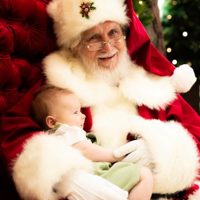 A baby in a green outfit is cradled in Santa's arms against his white fur-trimmed suit
