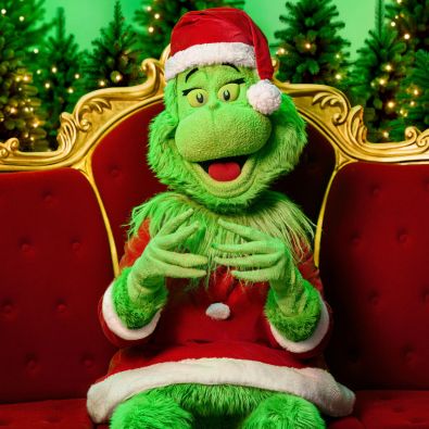 The Grinch in a Santa suit sits cheekily on a red velvet and gold throne, surrounded by lit trees.