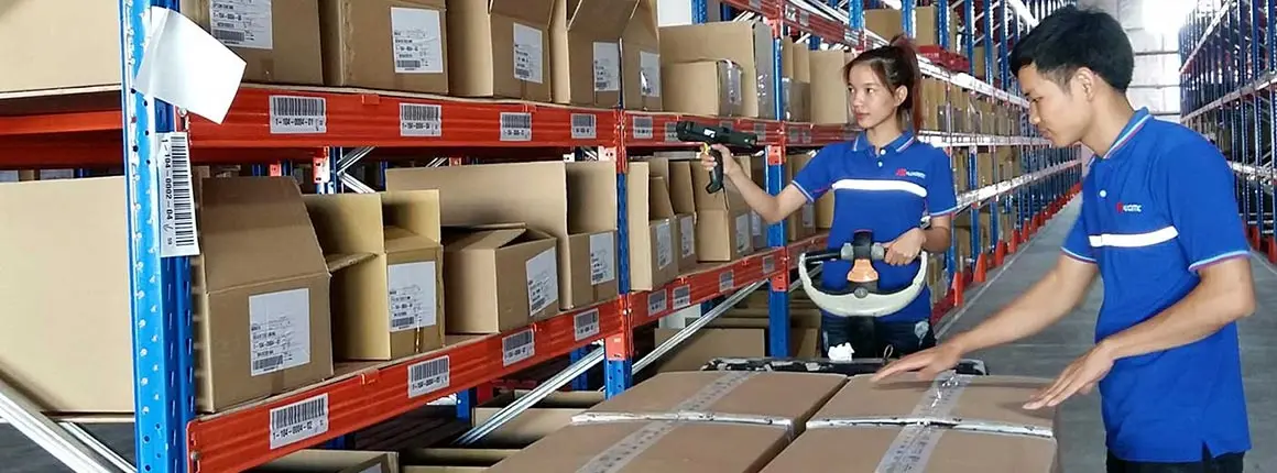 A photograph inside a warehouse or distribution center. A woman in a blue and white uniform is scanning a barcode on a box on a shelf using a handheld scanner. A man, also in a blue and white uniform, is standing next to her, inspecting or organizing boxes on a roller conveyor. The shelves behind them are full of various-sized cardboard boxes, many with visible barcodes and labels.