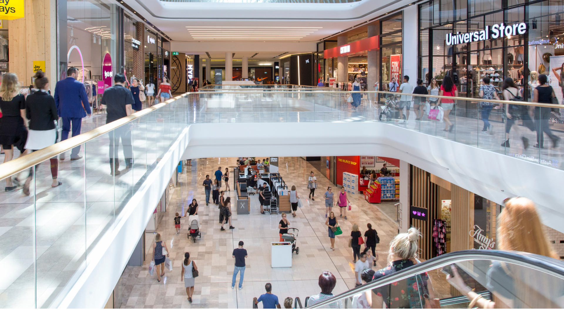 A busy, multi-level modern shopping mall with people walking on the concourses and riding an escalator
